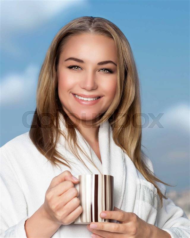 Portrait of cute smiling girl drinking tea on the balcony over blue sky background, wearing white bathrobe and having breakfast, with pleasure relaxing at home, stock photo
