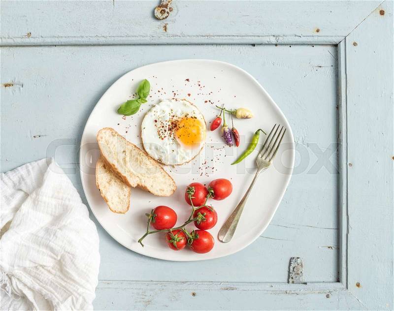 Breakfast set. Fried egg, bread slices, cherry tomatoes, hot peppers and herbs on white ceramic plate over light blue wooden backdrop, top view, stock photo