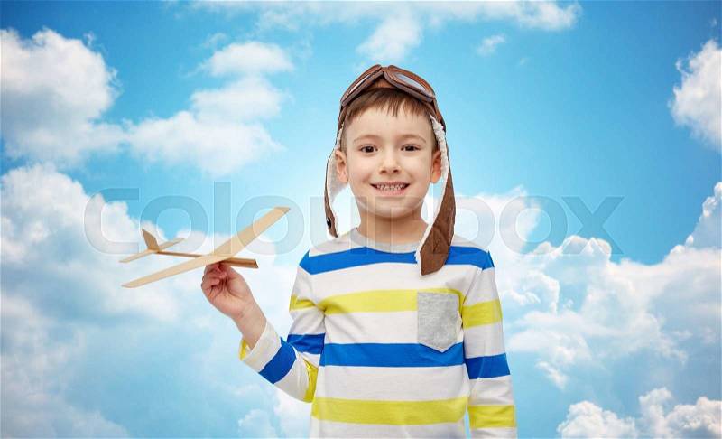 Childhood, fashion and people concept - happy smiling little boy in aviator hat playing with wooden airplane over blue sky and clouds background, stock photo