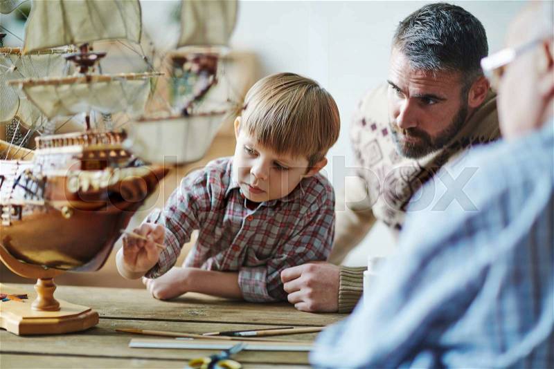 Adorable youngster painting toy ship with his father and grandfather near by, stock photo