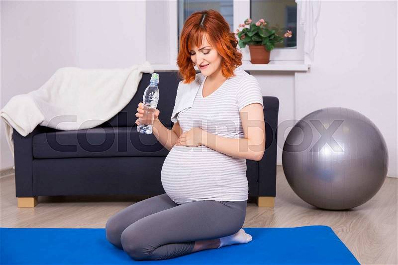 Young happy pregnant woman relaxing and drinking water after fitness training at home, stock photo
