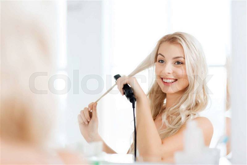 Beauty, hairstyle, morning and people concept - smiling young woman with styling iron straightening her hair and looking to mirror at home bathroom, stock photo