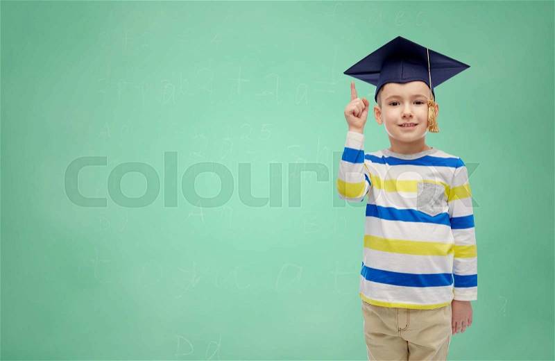 Childhood, school, education, learning and people concept - happy boy in bachelor hat or mortarboard pointing finger up over green chalk board background, stock photo