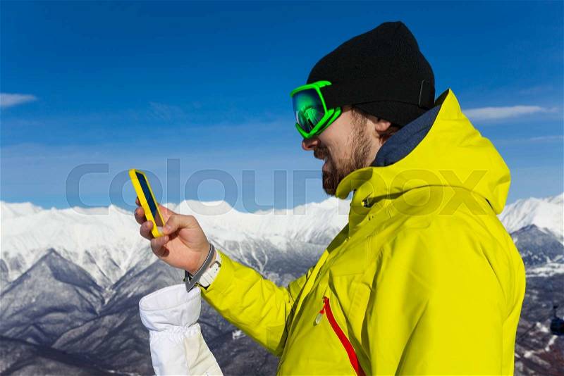 Man skier with beard and ski mask text holding phone standing on top of the mountain, stock photo