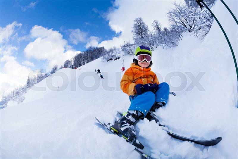 Little boy wearing helmet and ski mask sit in snow resting after skiing lessons, stock photo