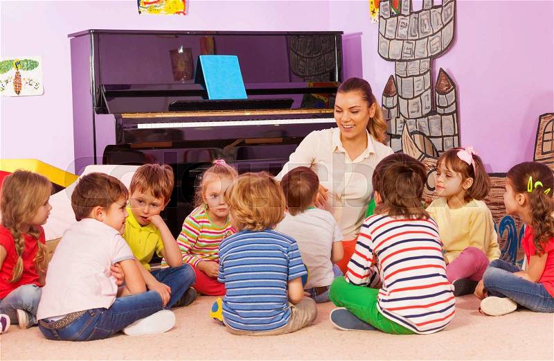 Group of kids sit with teacher and listening to story she tell in kindergarten room, stock photo