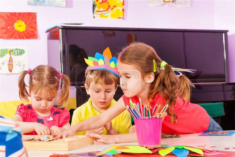 Little girl stretching for paper gluing in kindergarten class with other boys and girls , stock photo
