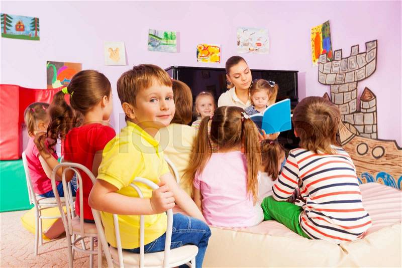 Boy portrait among large group of little kids sit in class and listen to teacher reading a book and telling stories, stock photo