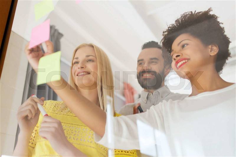 Business, startup, planning, management and people concept - happy international creative team writing on stickers at office glass board, stock photo