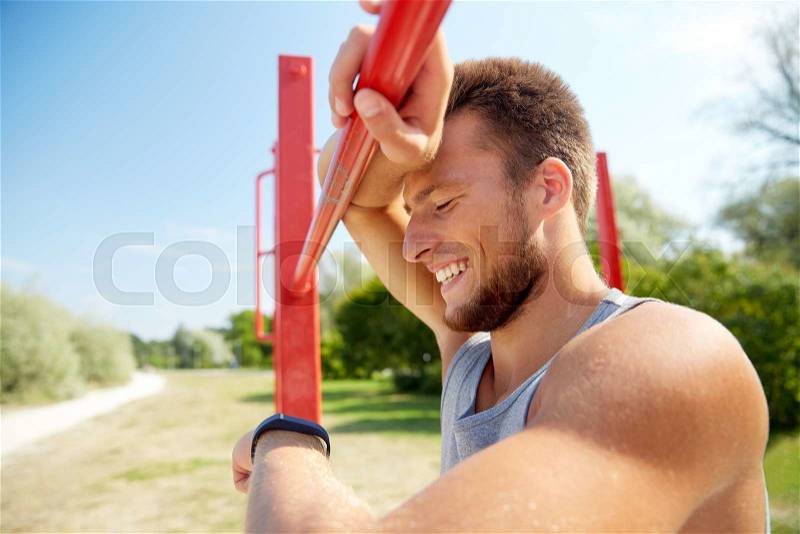Fitness, sport, training and lifestyle concept - happy young man looking at heart-rate watch bracelet and exercising on horizontal bar outdoors, stock photo