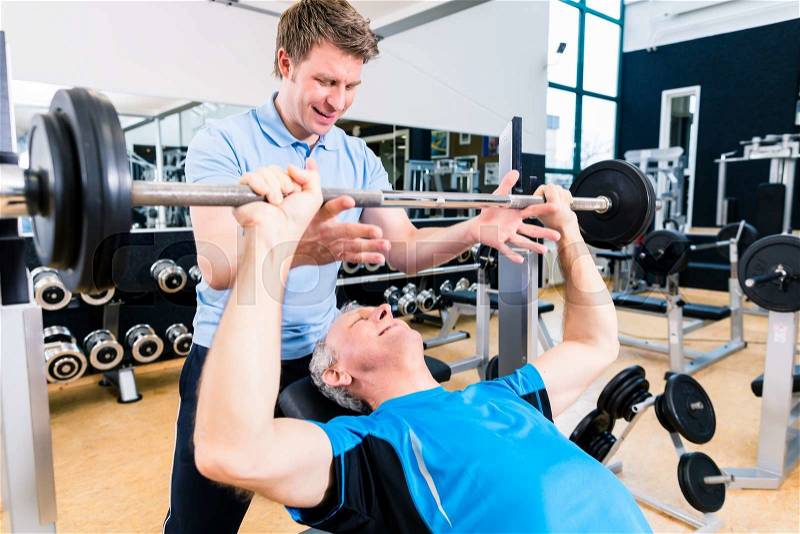 Trainer assisting senior man lifting barbell in gym to gain strength and fitness, stock photo