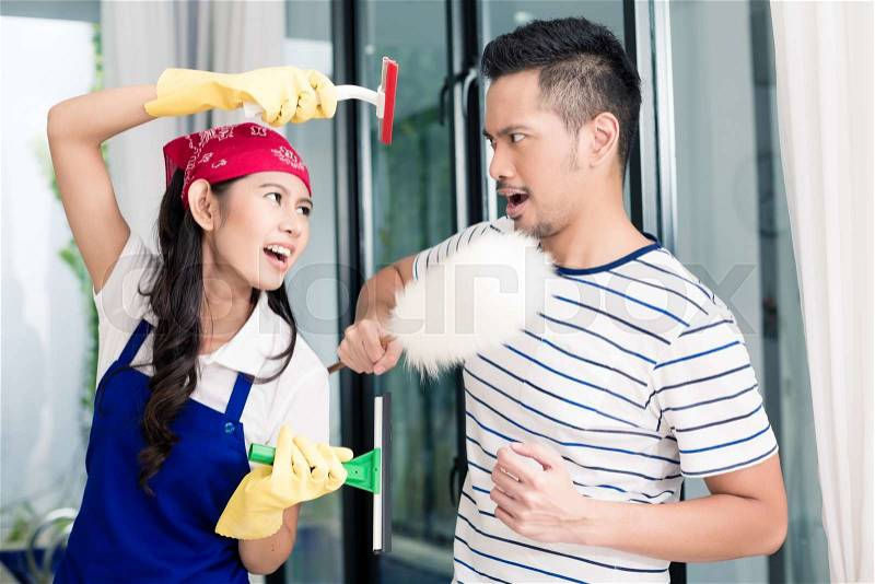 Asian woman and man having fun cleaning home staging a mock fight with chores utensils, stock photo