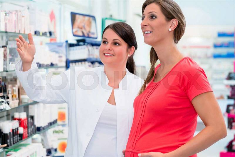 Pharmacist showing pregnant woman drugs in pharmacy or drug store, both women are standing in front of a shelf, stock photo