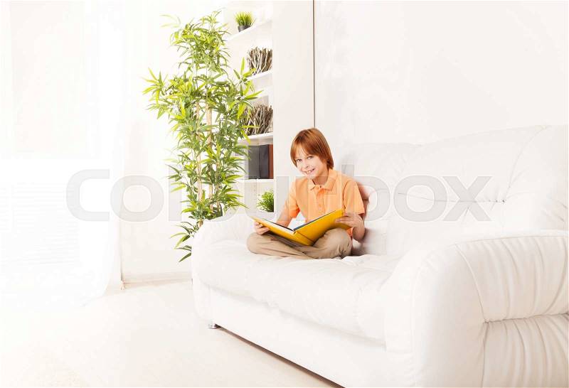 Smart smiling Caucasian boy portrait in interior sitting on the sofa at home and reading, stock photo