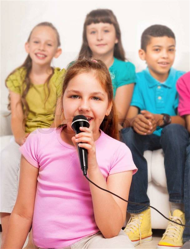 Close portrait happy beautiful girl sitting on the floor and singing to microphone with group of friends, boys and girls sitting on the coach on background, stock photo