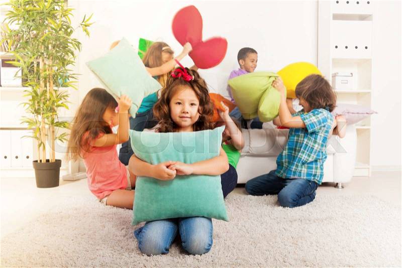 Happy smiling little Asian girl hugging pillow with large group of her friends pillow fighting on the background, stock photo