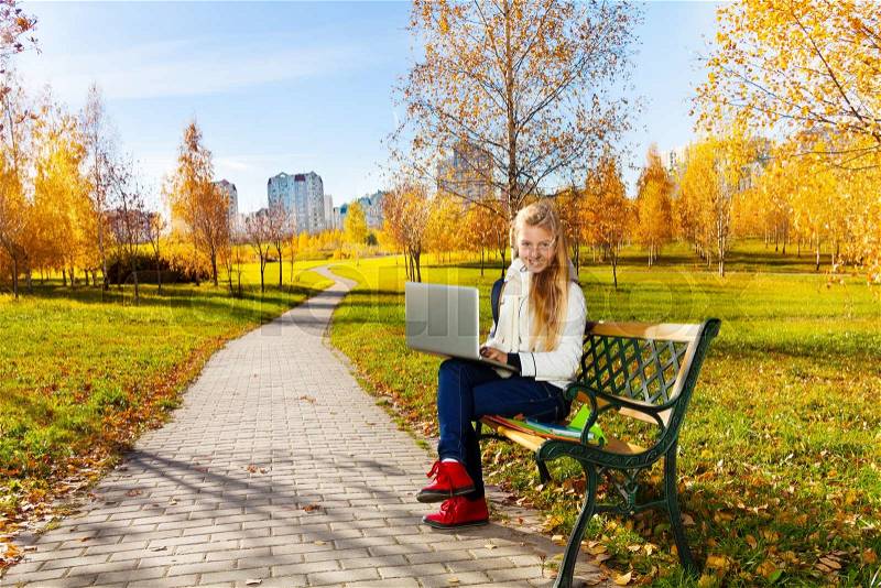 Happy blond teen girl with long hair sitting with laptop in the autumn park, stock photo