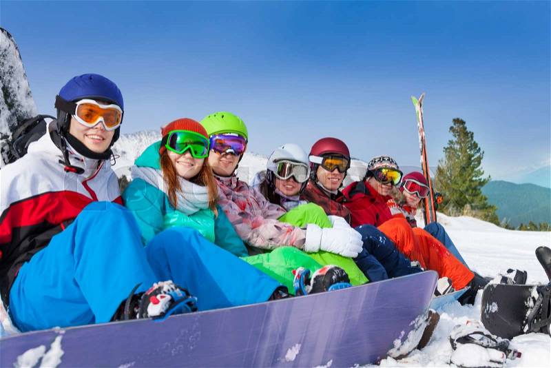 Portrait of seven smiling friends sitting in a row with snowboards and skis wearing goggles on the mountain background, stock photo