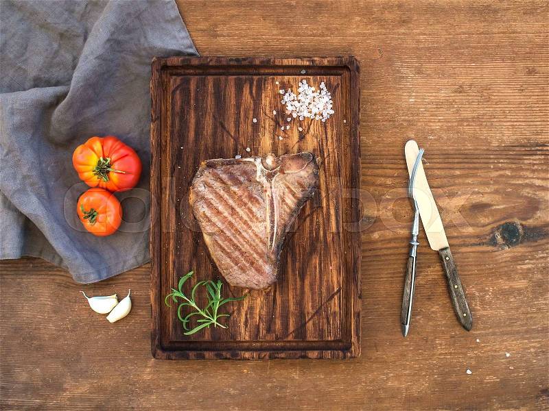 Cooked meat t-bone steak on serving board with garlic cloves, tomatoes, rosemary and spices over rustic wooden background, top view, stock photo