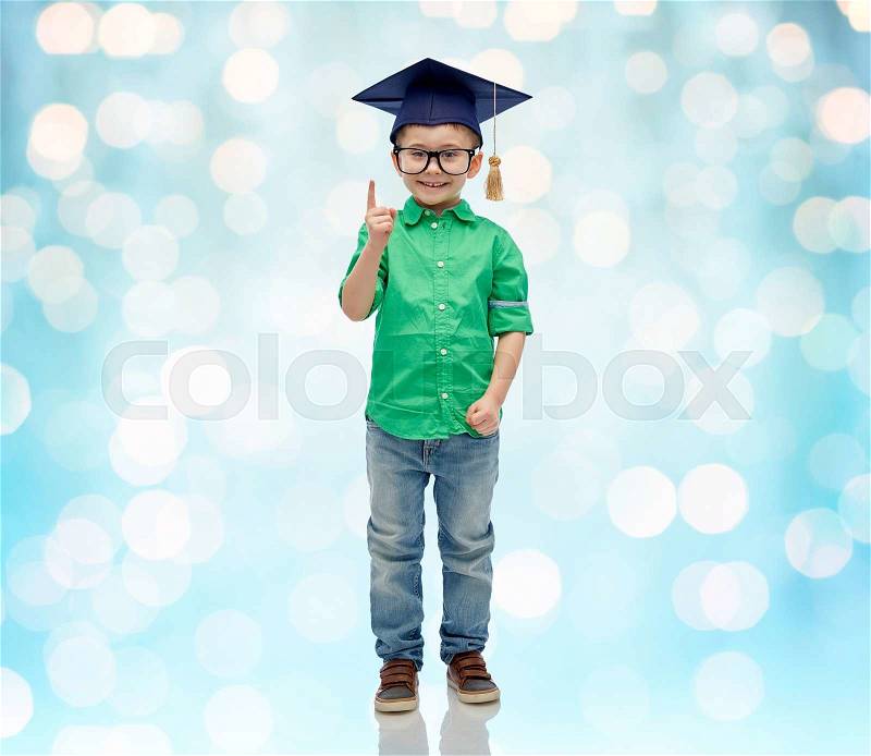 Childhood, school, education, knowledge and people concept - happy boy in bachelor hat or mortarboard and eyeglasses pointing finger up over blue holidays lights background, stock photo