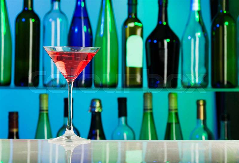 Glass with pink liquid stands on table of bar with green and blue bottles on background, stock photo