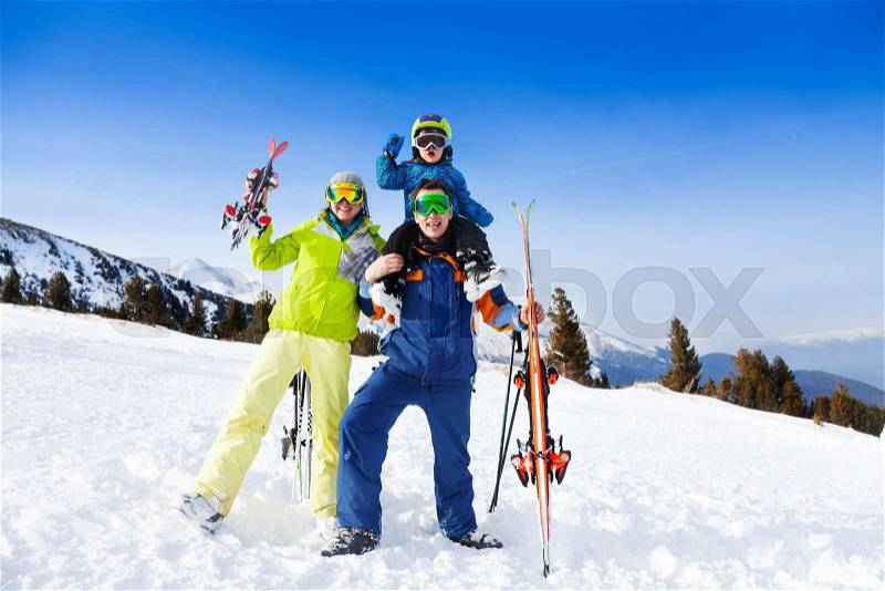 Mom, dad with child on his shoulders in ski masks holding ski on mountains background, stock photo