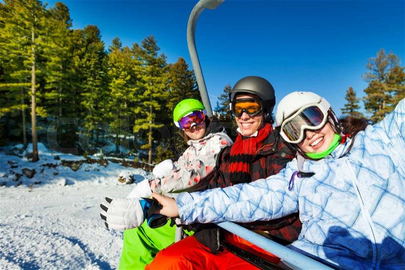 Three happy snowboarders in ski masks sitting together in elevator in mountains and forest area, stock photo