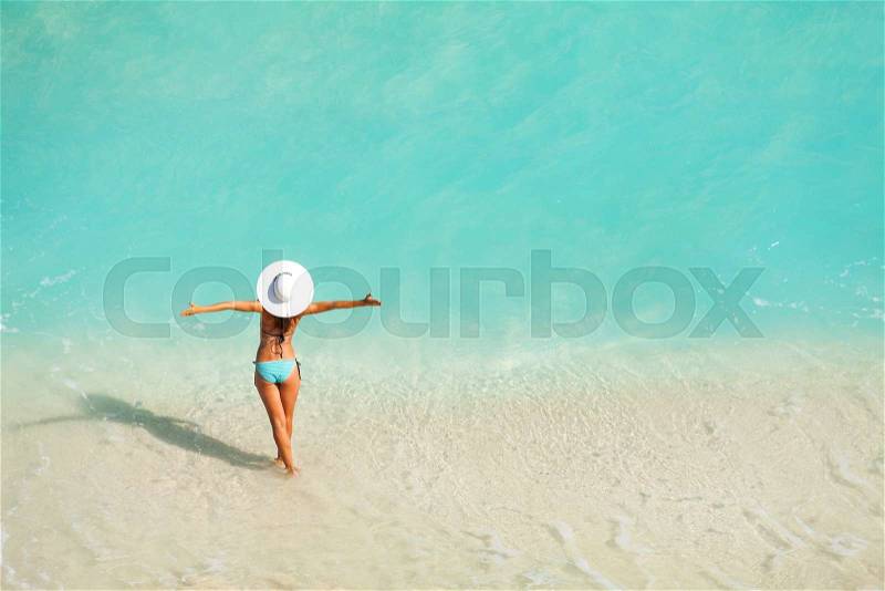 Top view of woman with white hat in striped bikini standing in ocean water on the sand of beach during holidays summer time, stock photo