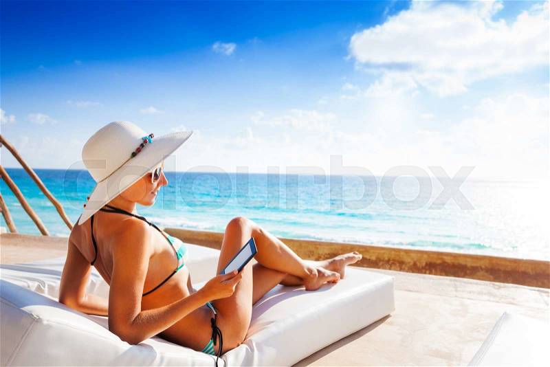 Beautiful young woman from back with white hat laying on white mattress and reading e-book with beautiful sea on background, stock photo
