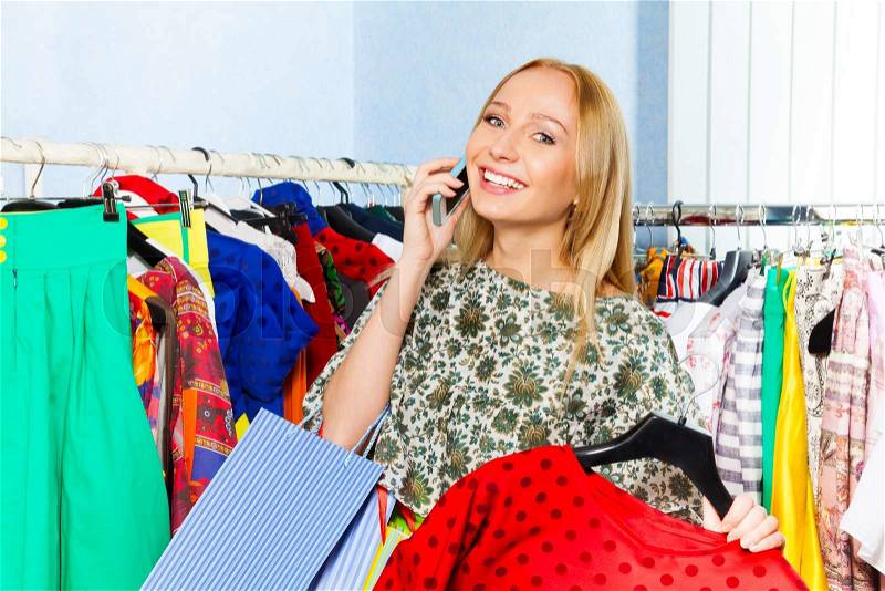 Young woman talking on mobile phone and holding chosen clothes in the shop, stock photo