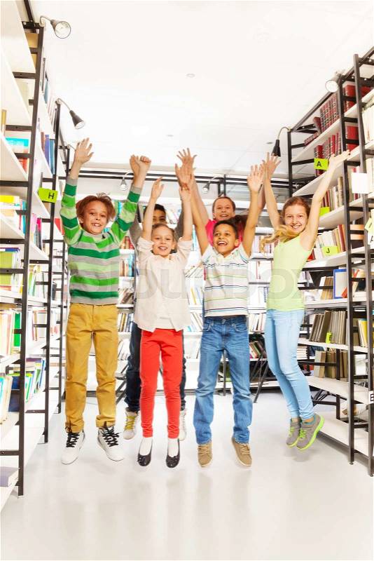 Happy kids jumping with hands up in the library and standing between bookshelves, stock photo