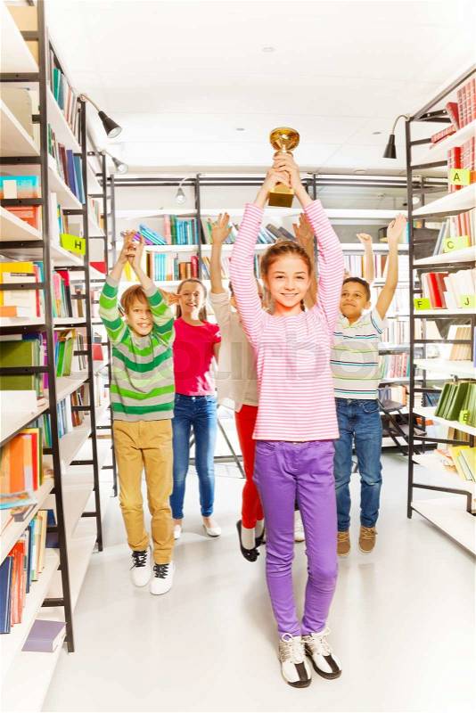 Happy girl holds golden cup above her head and other kids jumping behind in the library, stock photo