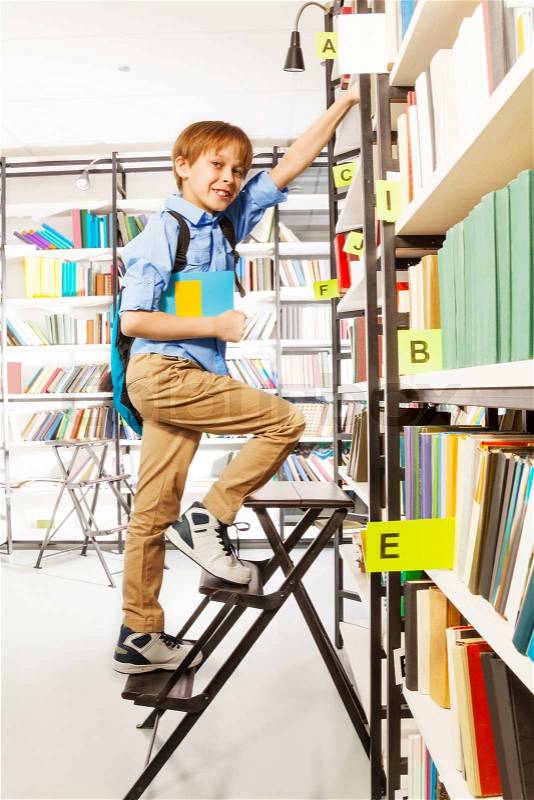 Boy climbing on step ladder in library with blue bag and colorful book, stock photo
