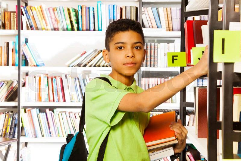 Serious boy looks straight and searches book on shelf in library, stock photo