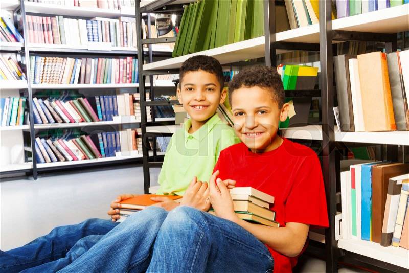 Two laughing boys sitting on the floor in library and holding two piles of books, stock photo