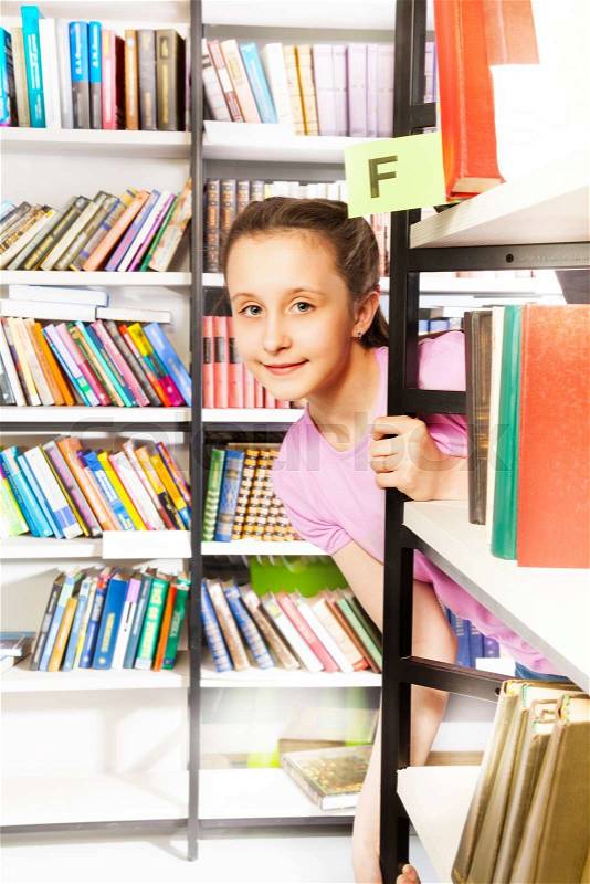 Cute girl is looking and standing behind the bookshelf and holding it in library, stock photo