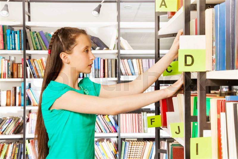 Schoolgirl with long hair searching books and holding exercise books on the bookshelf in library, stock photo