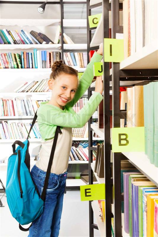 Schoolgirl with blue bag searching books and holding exercise books on the bookshelf in library, stock photo
