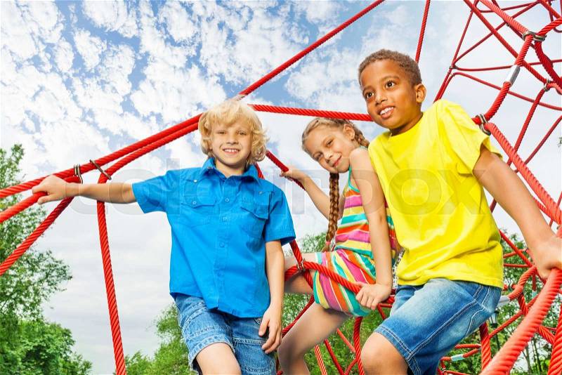 Two boys and girl sit on red ropes of playground and smile on sky background, stock photo