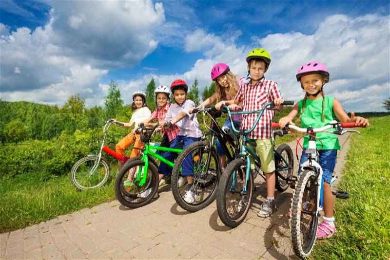 Children in row wearing colorful bike helmets holding bike handle-bars and are ready to ride their bikes in summer, stock photo