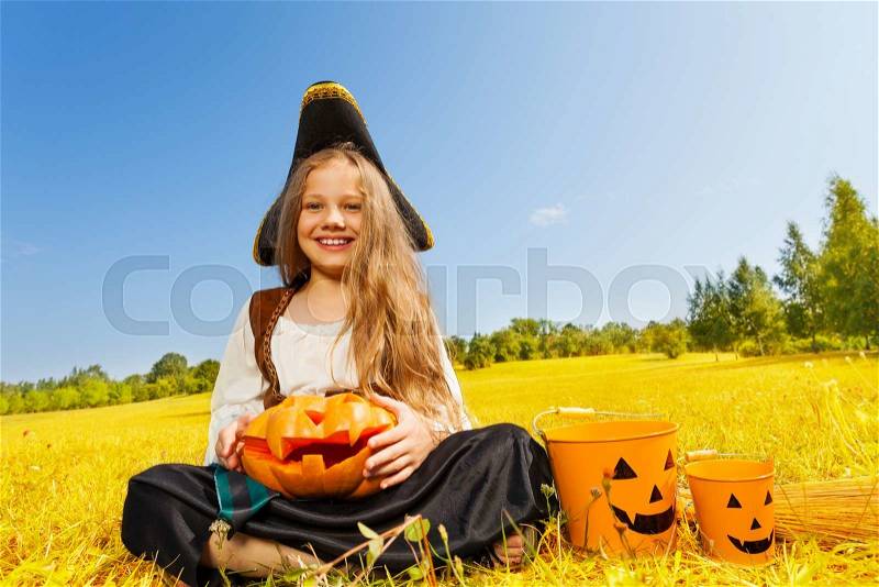 Halloween girl in costume of a pirate sitting on yellow grass with pumpkin and pail near her, stock photo