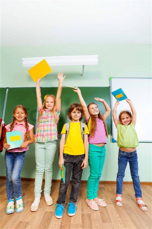 Happy pupils hold textbooks up in the air stand close together near blackboard in school, stock photo