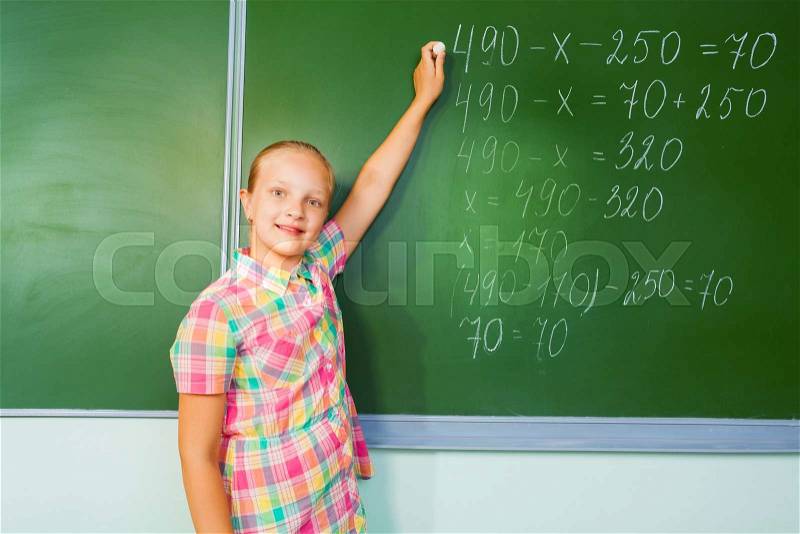Beautiful girl with chalk in her hand writing mathematics equation looking straight in front of blackboard during mathematics lesson, stock photo