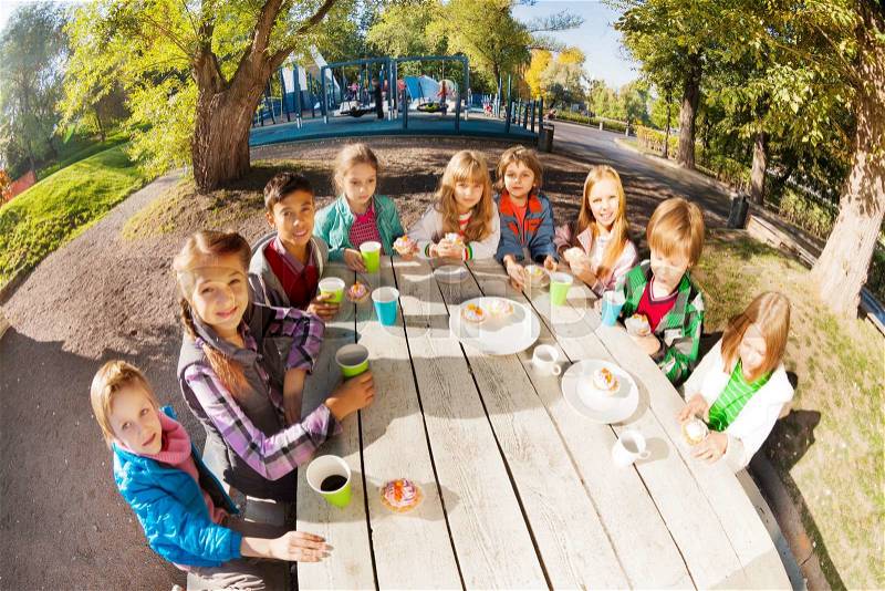 Wide angle lens view of international children drinking tea from colorful cups and eating cupcakes sitting at white wooden table outside during beautiful sunny day with green trees on background, stock photo