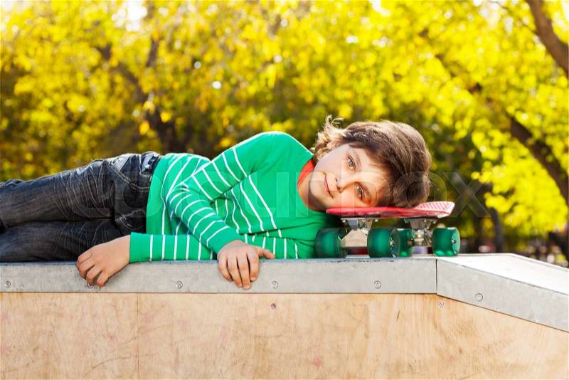 Small boy in green sweater laying on red skateboard during autumn sunny day on stoned ground, stock photo