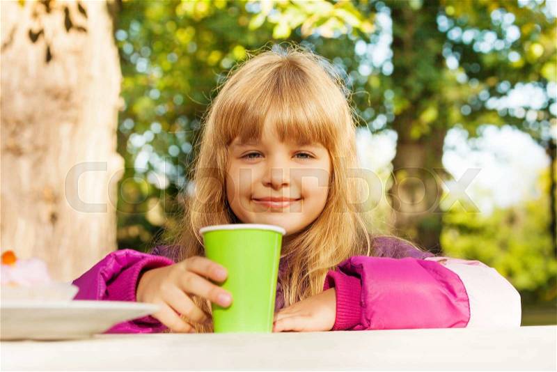 Cute small blond girl with green cup sitting at the table and drinking tea in park, stock photo