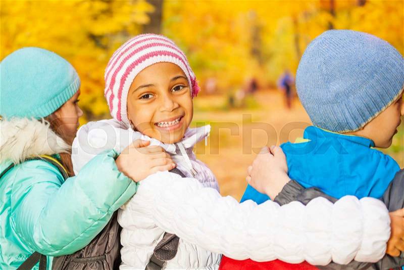 Happy children standing close with arms on shoulders of each other wearing rucksacks in the autumn forest during daytime, view from back with African girl turning back, stock photo