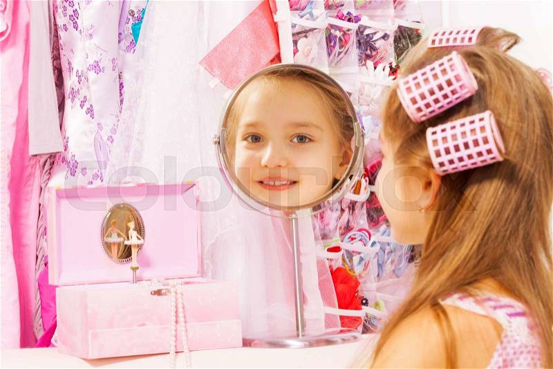 Beautiful small girl reflecting in round mirror with hair-curlers on her head while sitting turned back with accessories on background, stock photo