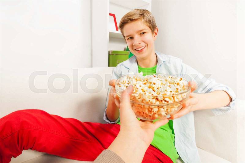 Smiling boy holds popcorn bowl from someone\'s hand on white sofa at home, stock photo