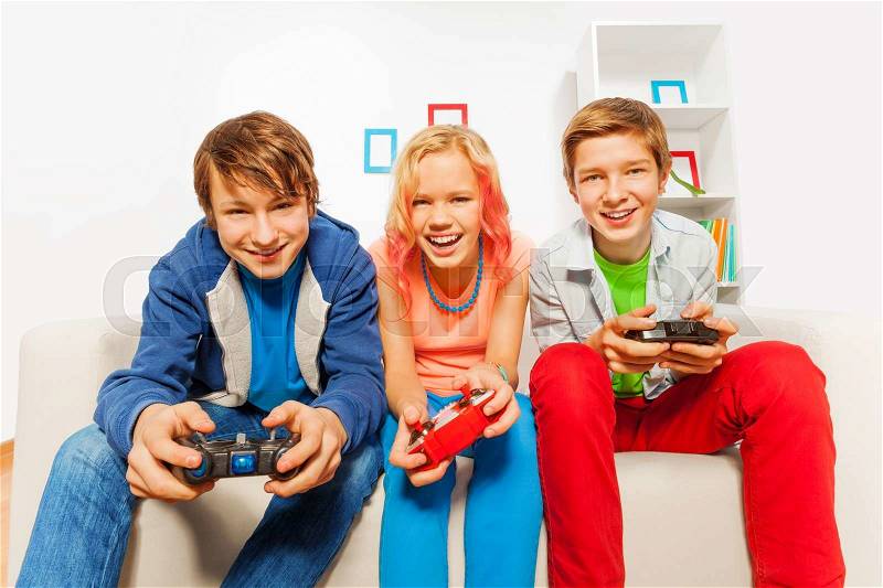 Happy teens holding joysticks and playing game console sitting on the sofa at home, stock photo